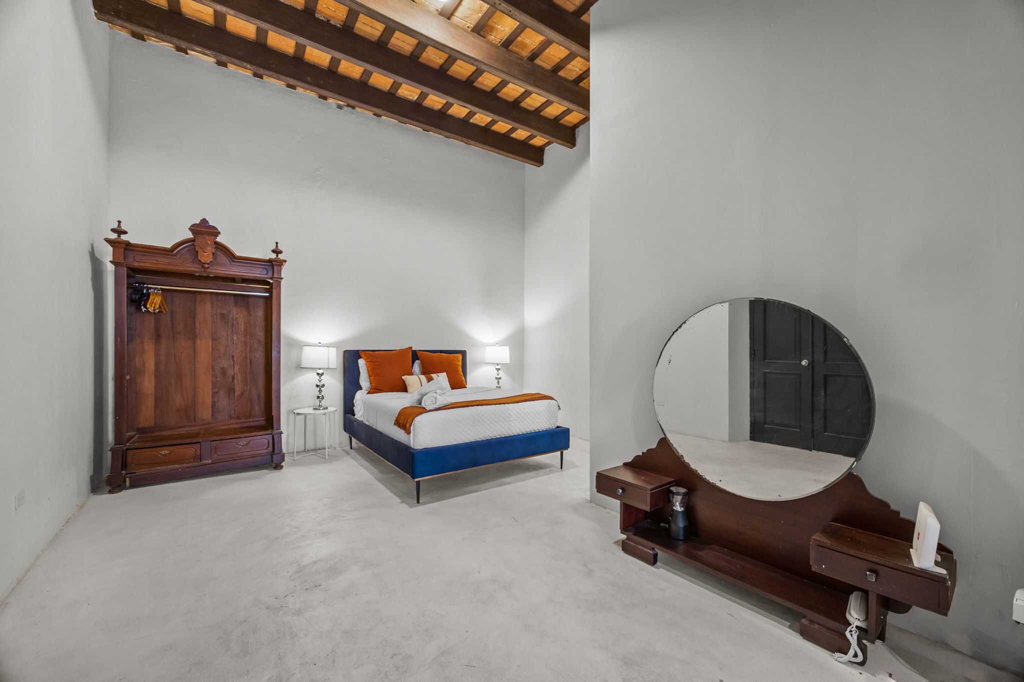 A bedroom with a bed, a wooden wardrobe, a round mirror, and a coffee machine.