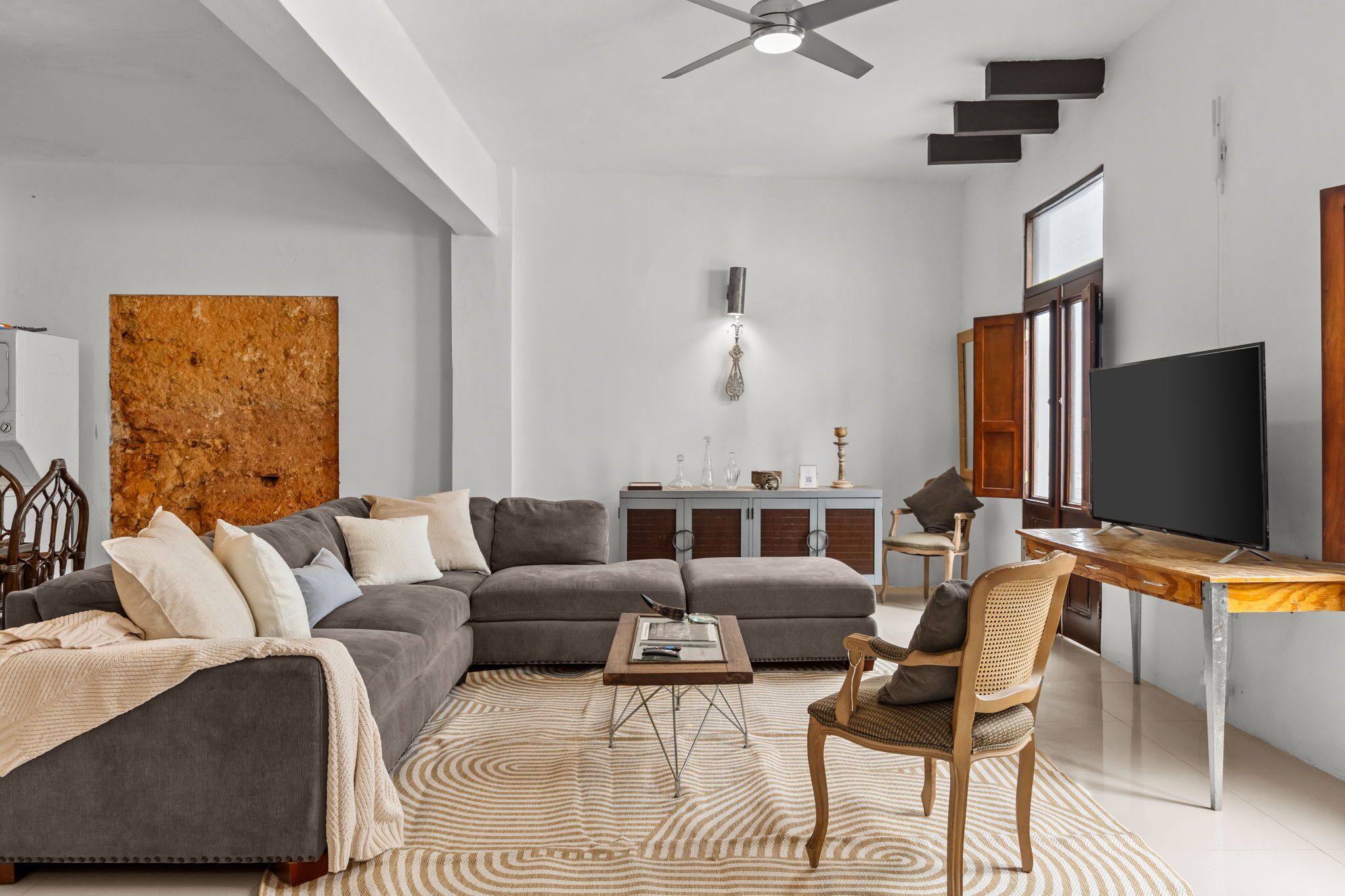 A modern living room with a sectional sofa, a wooden coffee table, a rattan chair, a television, and a decorative wall piece.