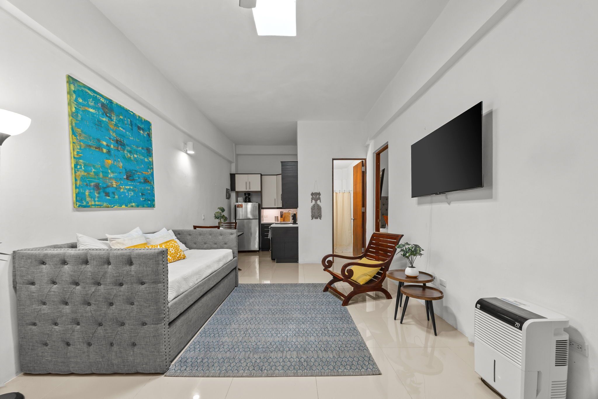 A modern apartment with a grey sofa, a dining table, a kitchen area, and a TV mounted on the wall.