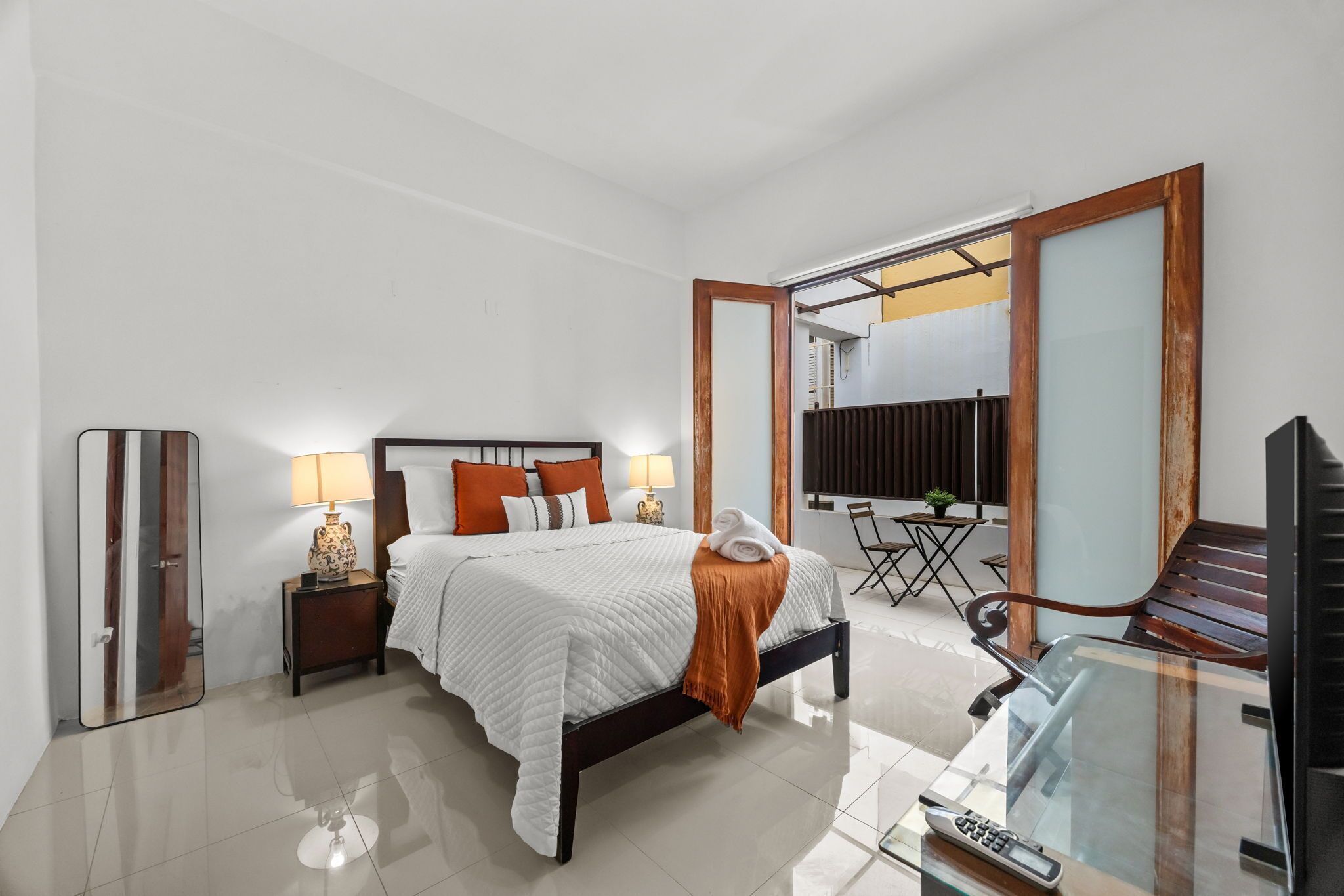 Un dormitorio con cama, mesitas de noche, un espejo, televisor, escritorio con silla y balcón con vistas.