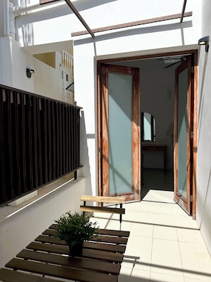 A narrow outdoor corridor with a wooden bench and a potted plant, leading to a room with a mirror and a lamp.