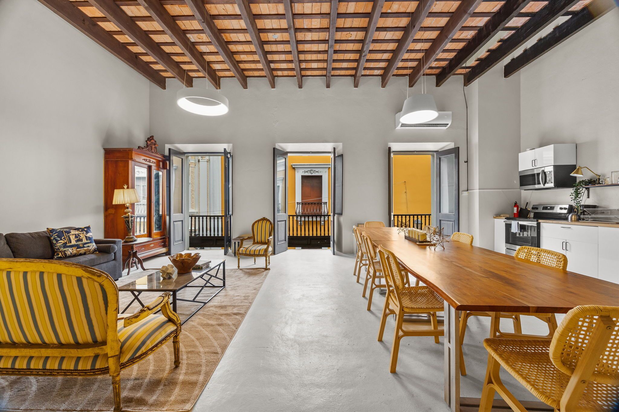 A spacious living area with a long wooden dining table, yellow-striped chairs, and a modern kitchen with stainless steel appliances.