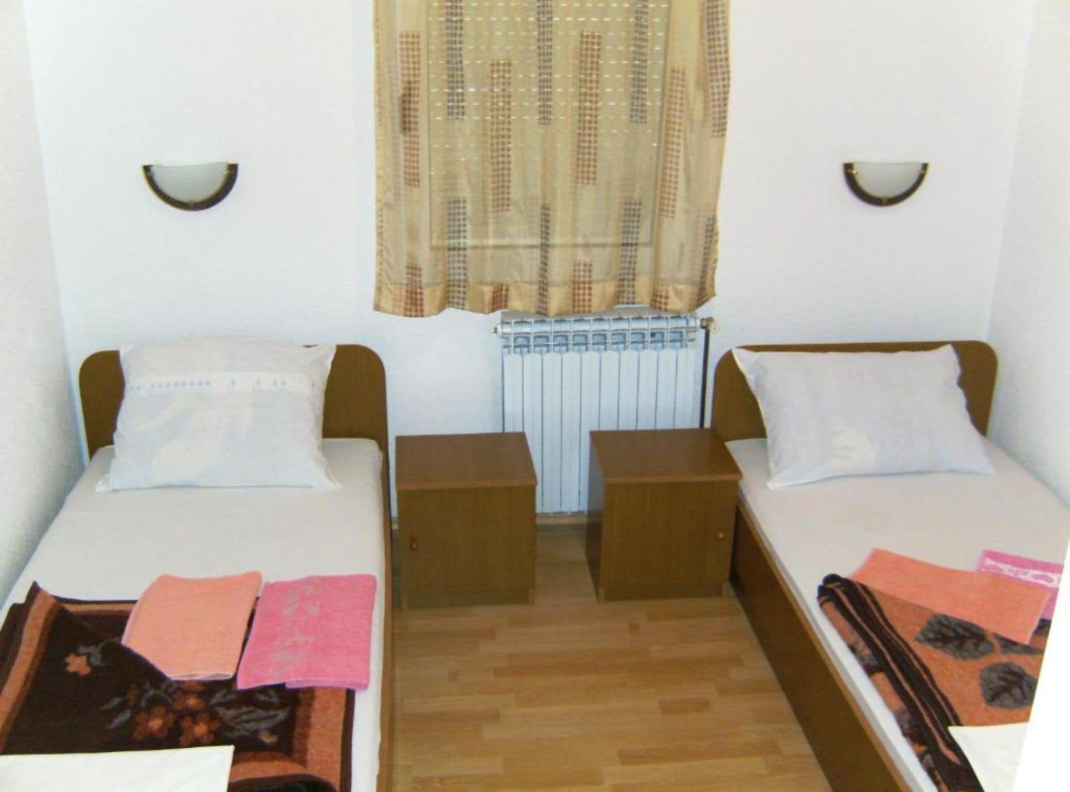 Quarto pequeno e limpo com duas camas individuais, chão de madeira, radiador e janela com cortina estampada.