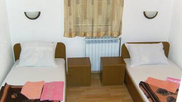 Quarto pequeno e limpo com duas camas individuais, chão de madeira, radiador e janela com cortina estampada.