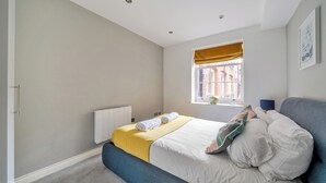 A bedroom with a bed, a window with a view, a radiator, and a blue bench. - Lace Market - 1-bed Apt - Sleeps 3 - City Centre (Nottingham)