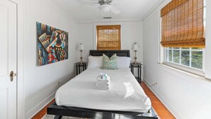Une chambre avec un grand lit, deux tables de chevet, un ventilateur de plafond, une fenêtre avec des stores en bambou et un tableau au mur.