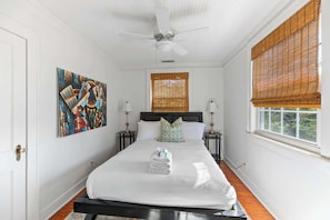 A bedroom with a large bed, two bedside tables, a ceiling fan, a window with bamboo blinds, and a painting on the wall. - Park Circle Gem, Near Everything + Daniels Is. (North Charleston)