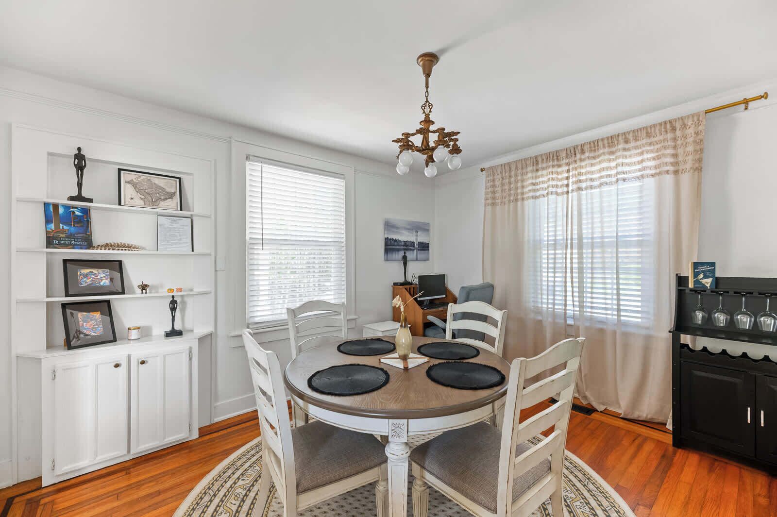 A dining area with a round table, white chairs, and a chandelier. There are shelves with framed pictures and a black cabinet with wine glasses.