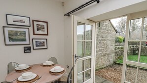 A dining area with a table set for four, framed pictures on the wall, and a stone wall outside. - Watermill Cottage (Chathill)