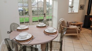 A dining area with a round table set for four, surrounded by chairs. There is a view of a garden through large windows, and a fireplace is visible in the background. - Watermill Cottage (Chathill)