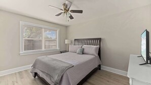 A bedroom with a bed, a ceiling fan, a window with blinds, a bedside table with a lamp, and a TV on a stand. - Stylish North Charleston Stay | Comfort + Convenience (North Charleston)