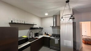 A compact kitchen with a stove, sink, and modern appliances. There are wine glasses on a shelf and a dining area with a table and chairs.