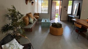 A spacious living area with a sofa set, a dining table, and a rocking chair. There is a large window providing natural light and a view of the outdoors.