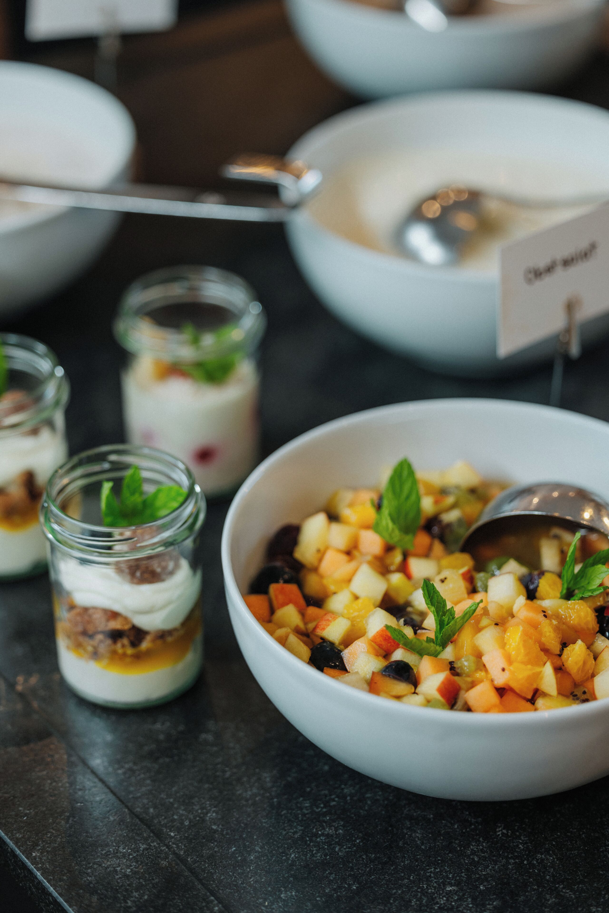 Un tazón de ensalada de frutas con adorno de menta, una cuchara y un frasco pequeño de yogur con nueces.