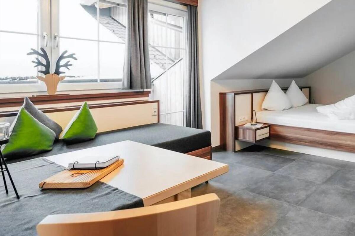 A modern hotel room with a desk, a bed with a wooden headboard, and a large window with curtains.