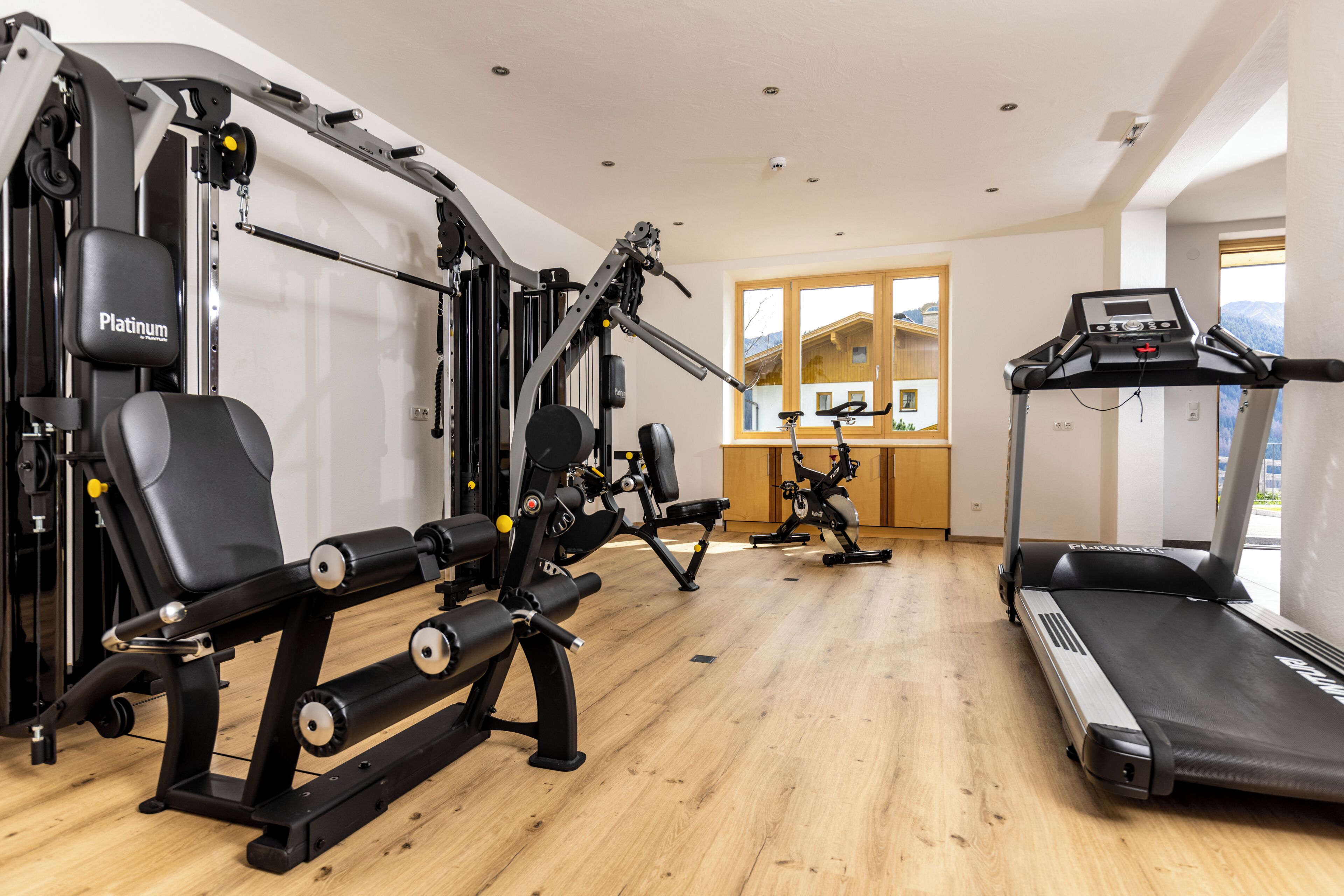 Un gimnasio con cinta de correr, máquinas de pesas y un ventanal con vistas a casas.