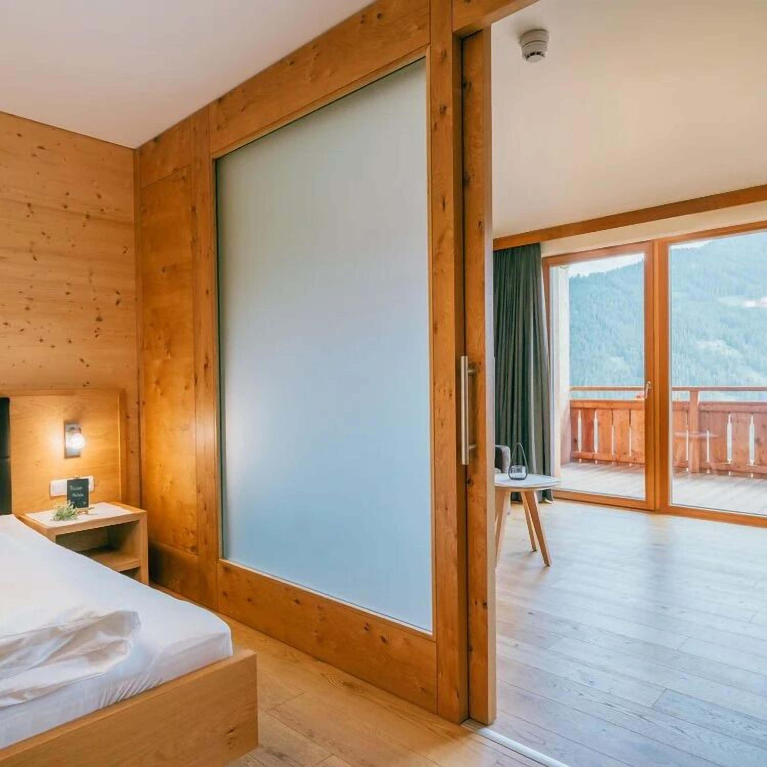 Una stanza in legno con un letto, un comodino e una porta scorrevole che dà su un balcone con vista su una montagna.