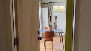 Une porte ouverte donne sur un coin repas, où se trouvent une table et des chaises, une plante en pot et une fenêtre avec des rideaux.