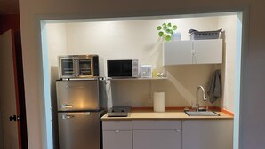 Une petite cuisine équipée d’un réfrigérateur, d’un four à micro-ondes et d’un four grille-pain. On y trouve un évier avec un robinet et un petit plan de travail. Une serviette est accrochée au mur, et une plante est posée sur une étagère au-dessus de l’évier.