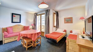 Una habitación acogedora con una cama de madera, una mesita y una silla. Hay una ventana con cortinas, un cuadro enmarcado en la pared y una lámpara.