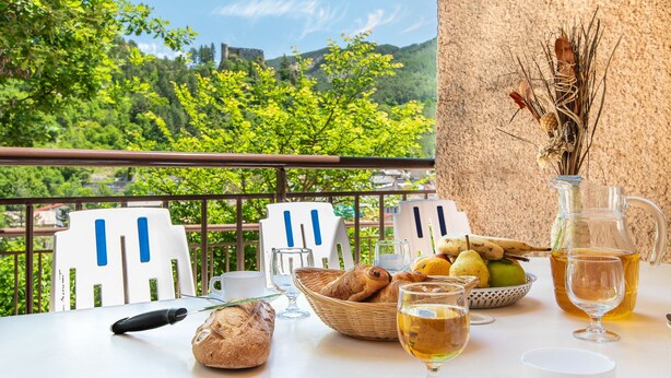 Una mesa con comida y bebidas, una cesta de pan, una jarra de té, y de fondo, vistas a la vegetación y un castillo.