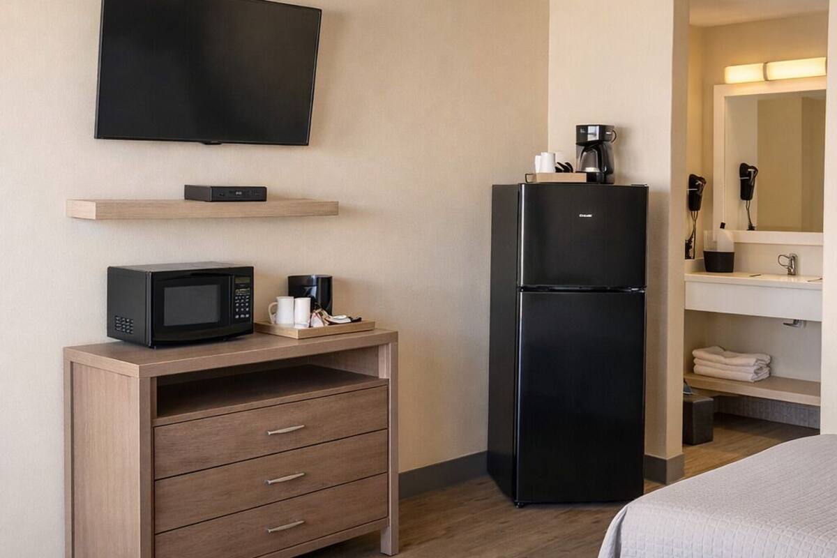 A hotel room with a bed, a desk with a microwave, a refrigerator, a TV, and a bathroom with a mirror and sink.