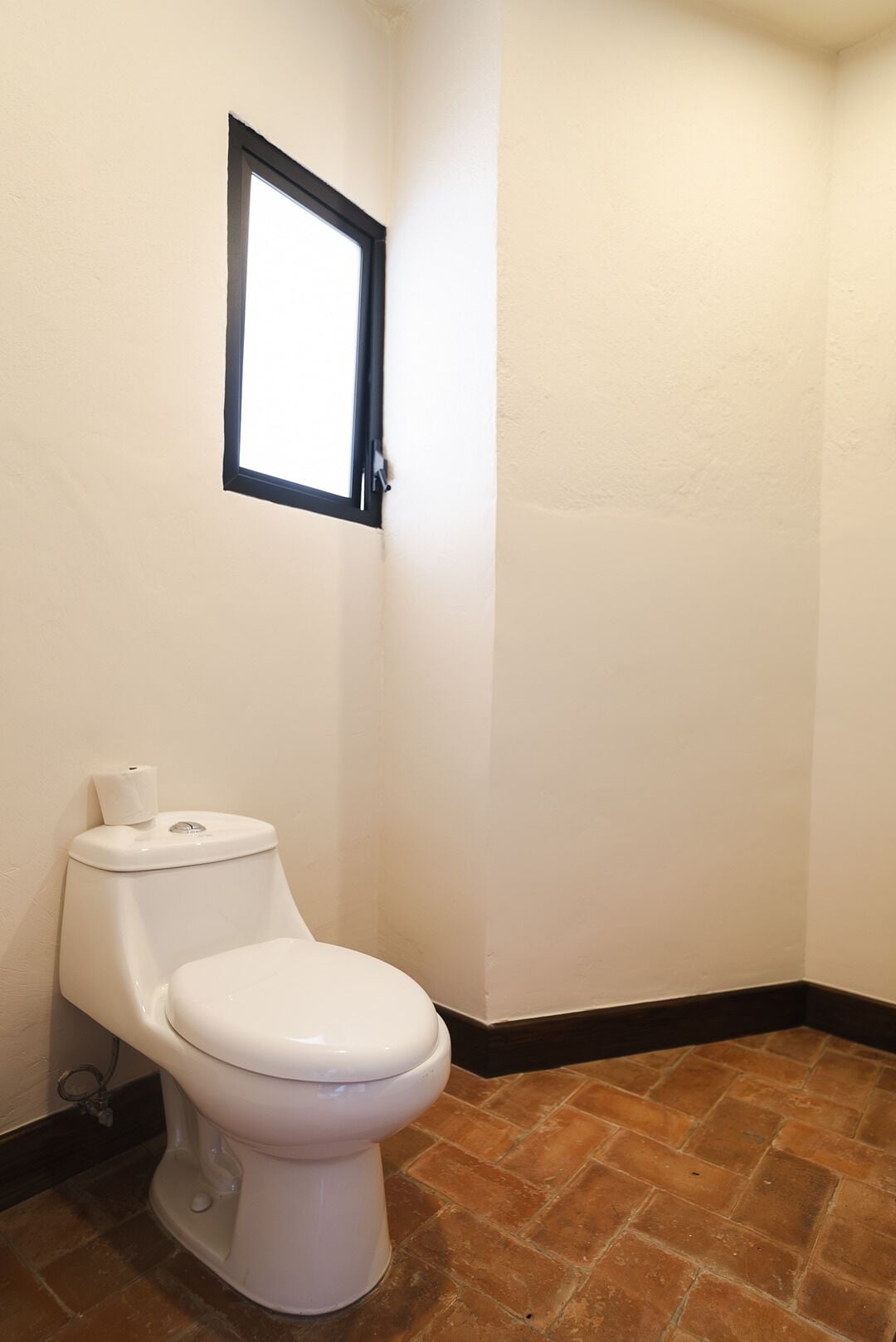 Ein Badezimmer mit Toilette, einem kleinen Fenster und braunen Fliesen.