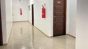 Un couloir aux murs blancs, doté d’un sol carrelé et d’une porte en bois sur laquelle est fixé un extincteur rouge.