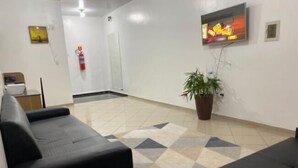 Un espace d’attente moderne avec un canapé noir, un tapis à motifs, un téléviseur fixé au mur et une plante en pot.