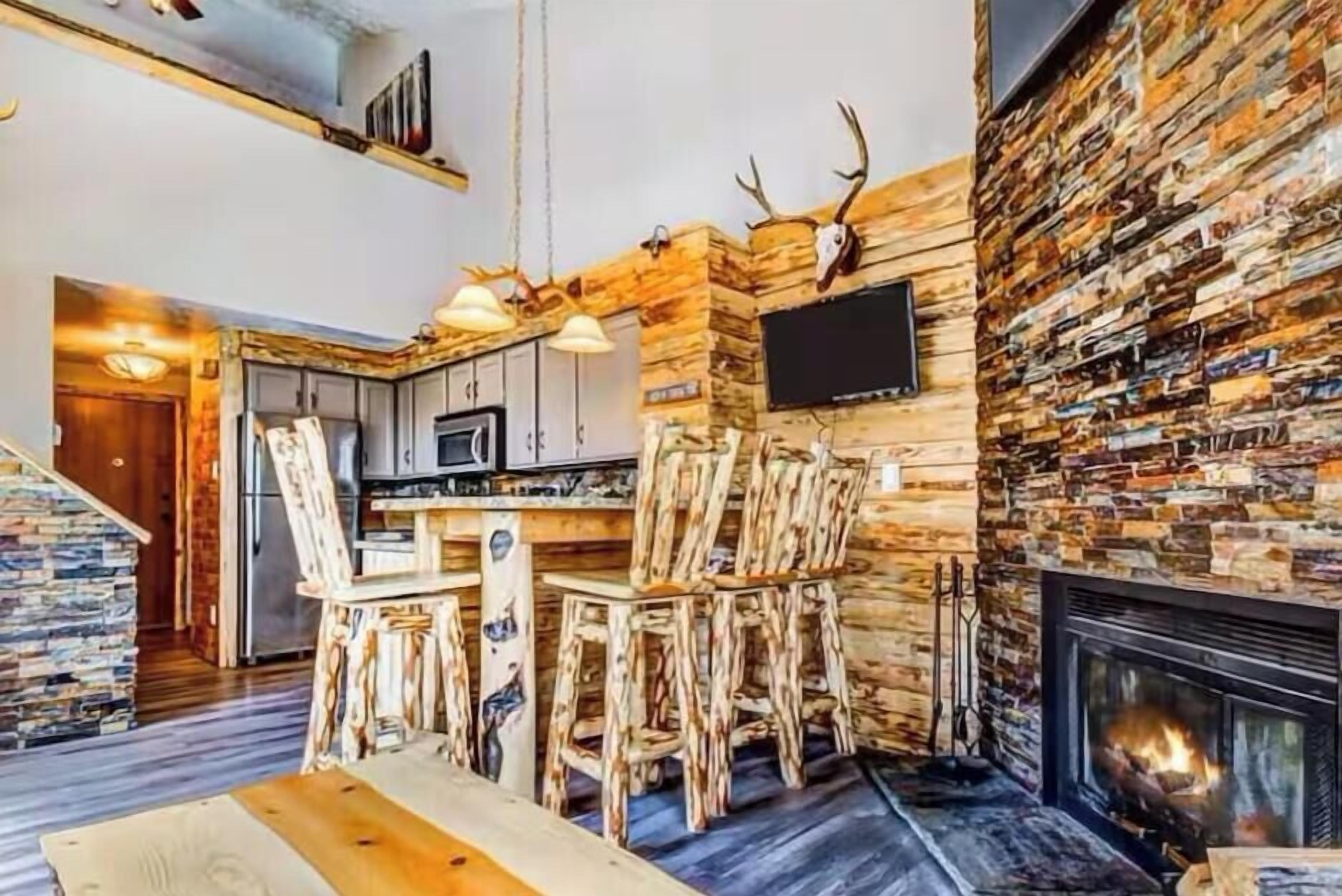 A rustic kitchen with a stone wall, a fireplace, and wooden chairs.
