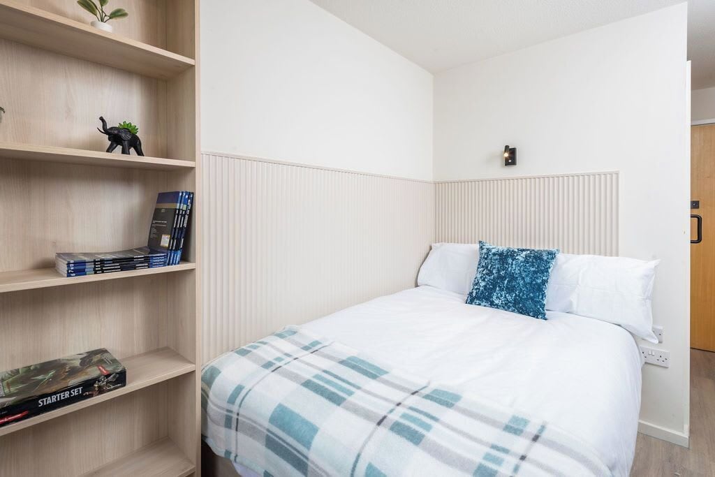 Un dormitorio ordenado con una cama, una estantería con libros y una figurita de dinosaurio, y un radiador.