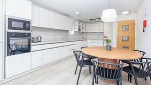 A modern kitchen with white cabinets, a built-in oven, and a round wooden dining table with black chairs. - Student Only The Railyard (Liverpool)