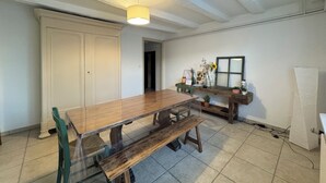 Un comedor con una mesa de madera, bancos y sillas. Se observa una puerta blanca, una ventana con vistas y una estantería con objetos decorativos.