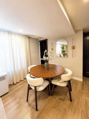 A dining area with a round wooden table, white cushioned chairs, a vase with white flowers, a mirror, and a dark door.