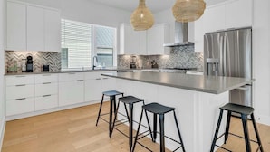 A modern kitchen with white cabinets, a large island with bar stools, a stainless steel refrigerator, and a herringbone tile backsplash. - Luxury Pool Retreat, Game Room, Sleeps 24 (Austin)