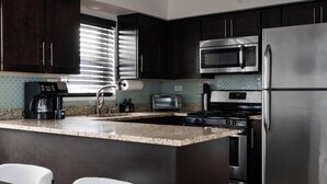 A modern kitchen with dark cabinets, stainless steel appliances, a granite countertop, and a window with blinds. - 2BR • Newly Furnished Modern Apt • Near O'Hare (Chicago)