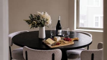 Un coin repas avec une table ronde, quatre chaises, un vase de fleurs, une bouteille de vin, des verres et une planche à fromages accompagnée de crackers et de fruits.