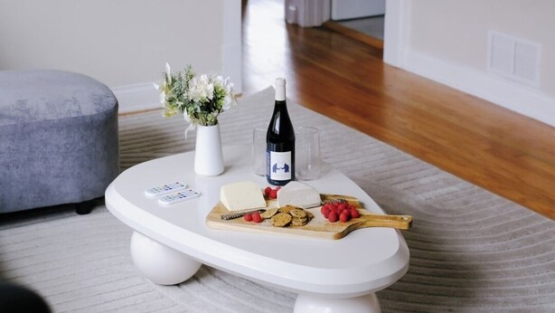 Ein modernes Wohnzimmer mit einem weißen Couchtisch, einer Blumenvase, einer Weinflasche und einem Käsebrett mit Snacks.