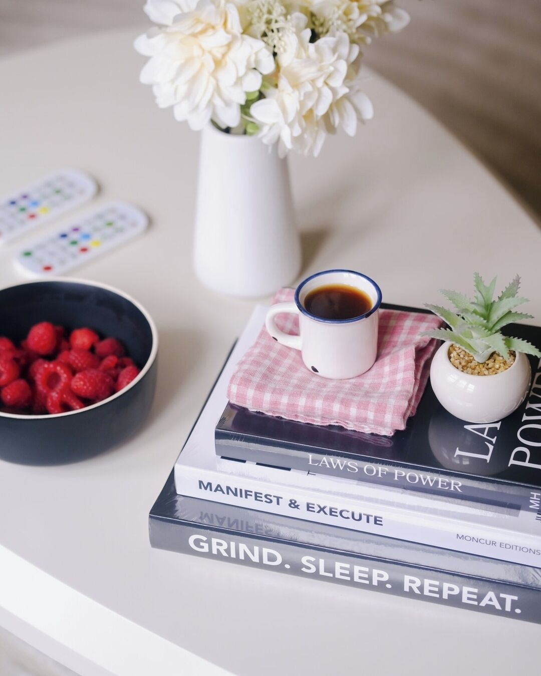 A table with a book titled "MANIFEST & EXECUTE," a cup of coffee, a bowl of raspberries, a small potted plant, and a vase with white flowers.