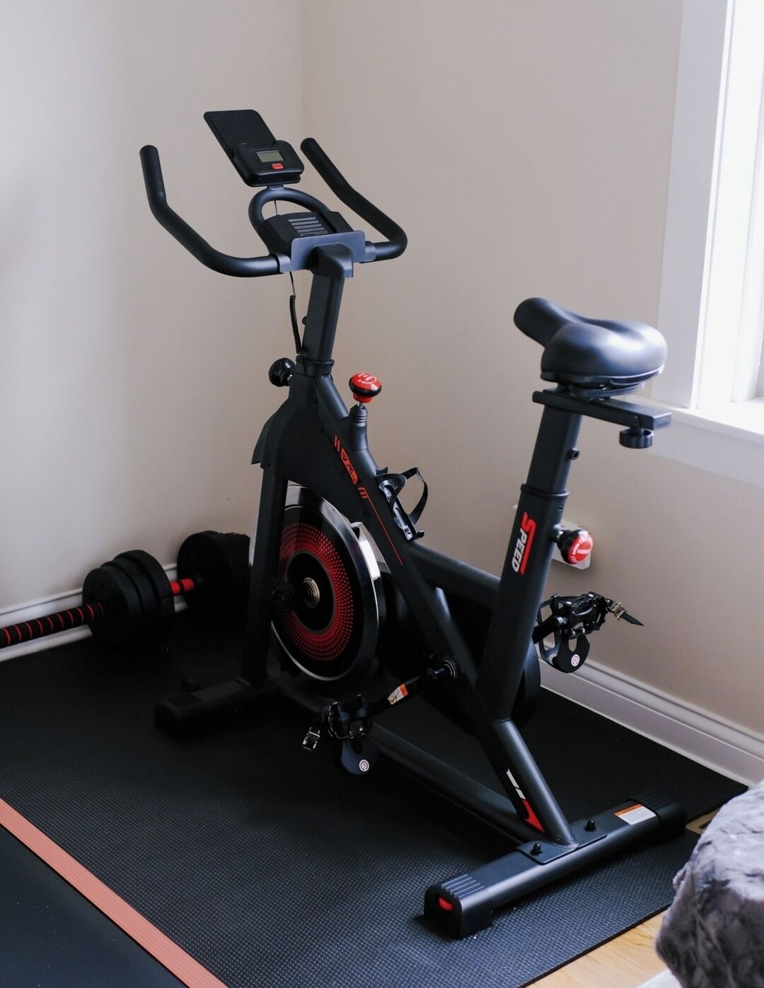 Een stilstaande fiets met een digitaal display, geplaatst op een mat in een kamer met een raam.