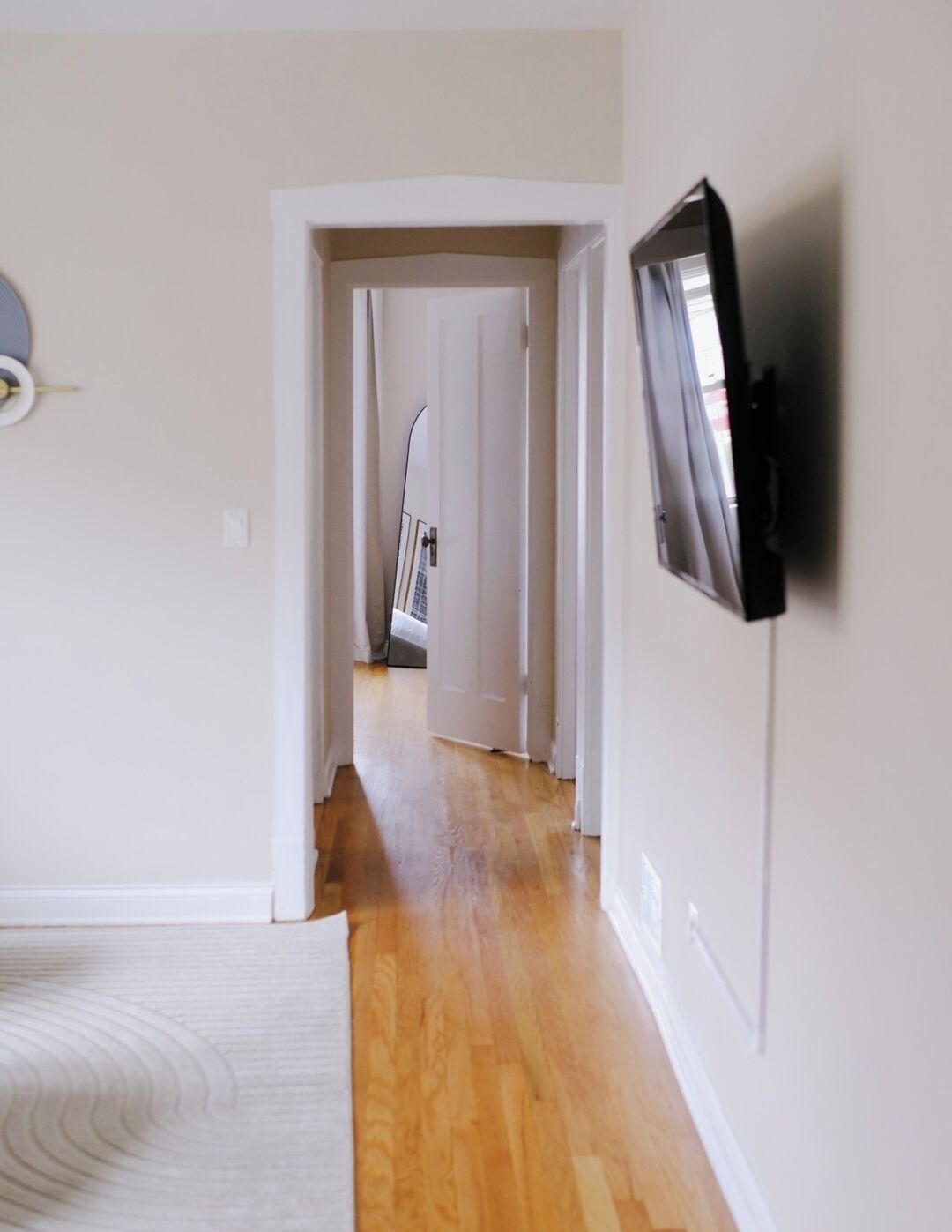 Un couloir au sol en bois, une porte blanche au fond et un téléviseur à écran plat fixé au mur.