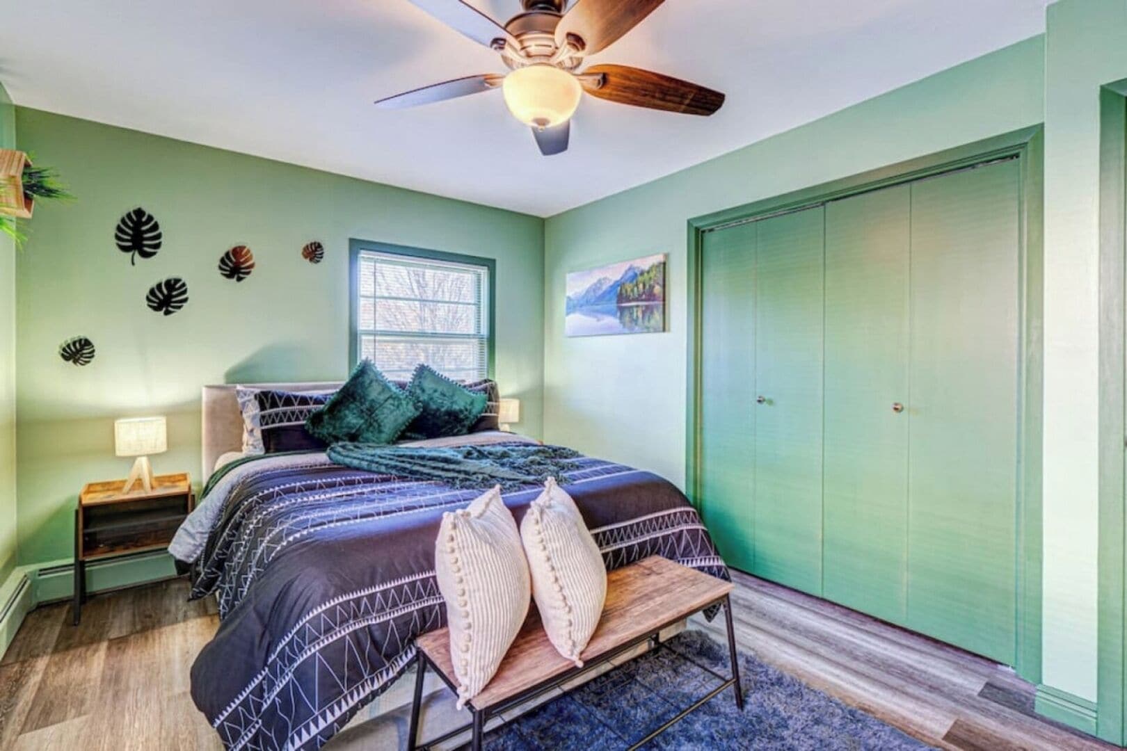 A bedroom with a bed, a nightstand, a bench, a ceiling fan, a window, and a green wardrobe.