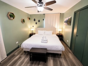 A hotel room with a bed, bedside tables, a ceiling fan, a window with curtains, and a door.