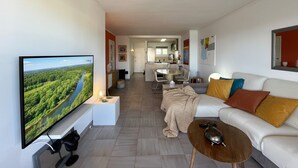 Ein modernes Wohnzimmer mit Flachbildfernseher, einer Couch, einem Couchtisch und einer Küche im Hintergrund.
