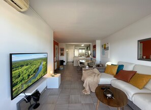 A modern living room with a flat-screen TV, a sofa, a coffee table, and a kitchen in the background. - Sea View Apartment Between Forest and Beach – Cala Vinyes (Badia de Palma)