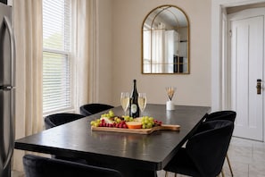 A dining area with a long table, black chairs, a fruit and cheese board, two glasses of white wine, and a mirror on the wall.