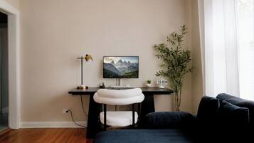 A room with a desk, a chair with a white cushion, a potted plant, and a large window.