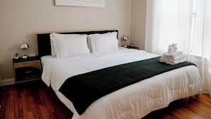 Ein ordentlich bezogenes Bett mit weißen Leinentüchern, einem schwarzen Kopfteil und einer dunkelgrünen Tagesdecke. Darüber an der Wand hängt ein gerahmtes Bild, und neben dem Bett liegt ein Stapel weißer Handtücher.