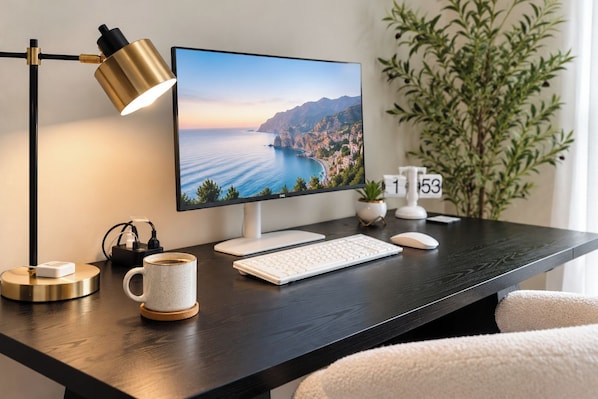A desk with a computer monitor, keyboard, mouse, a lamp, a mug, a plant, and a clock.