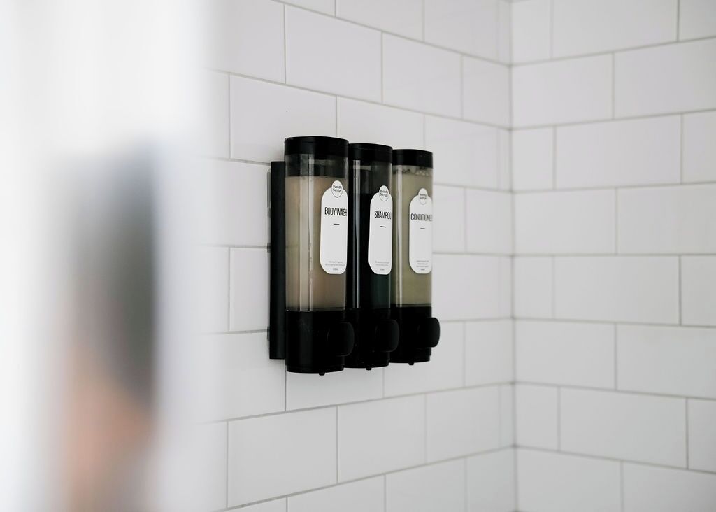 A wall-mounted dispenser with three bottles labeled 'BODY WASH,' 'SHAMPOO,' and 'CONDITIONER.'